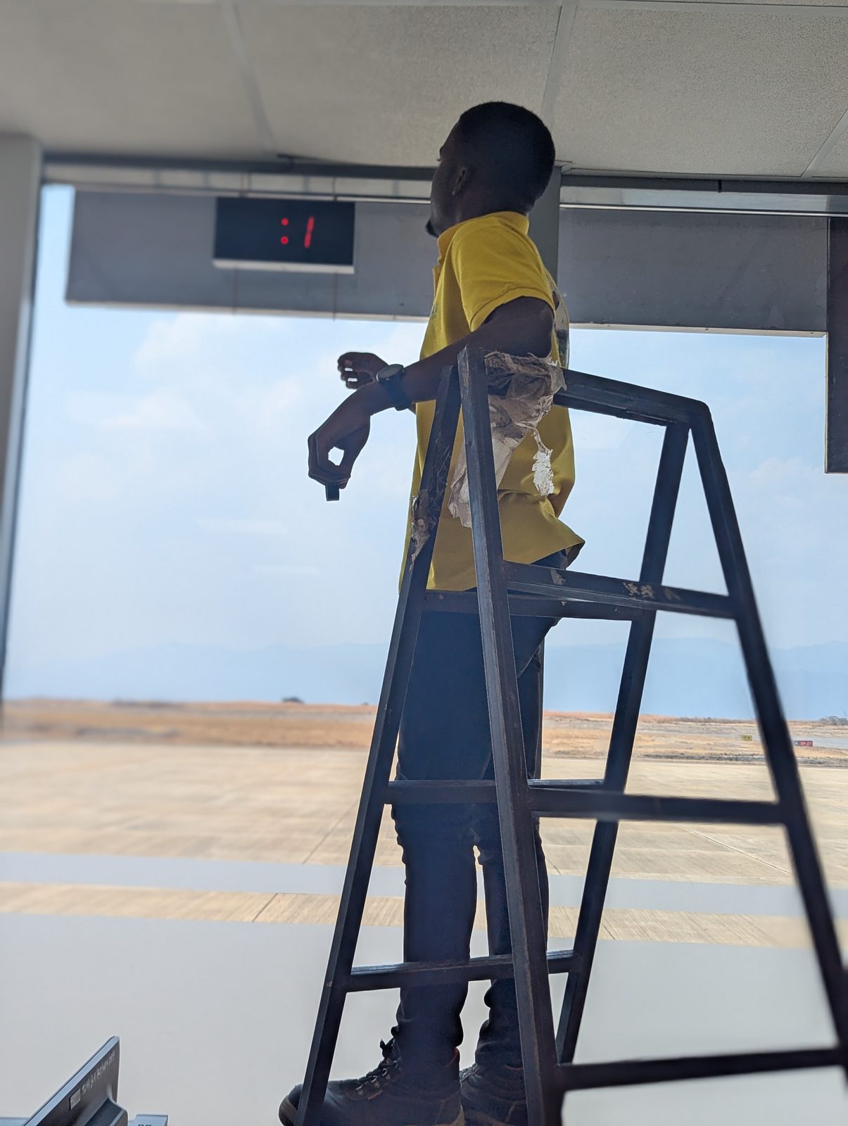 Lametronics engineer installing the digital clock at Songwe Airport, with the runway visible through the window behind him